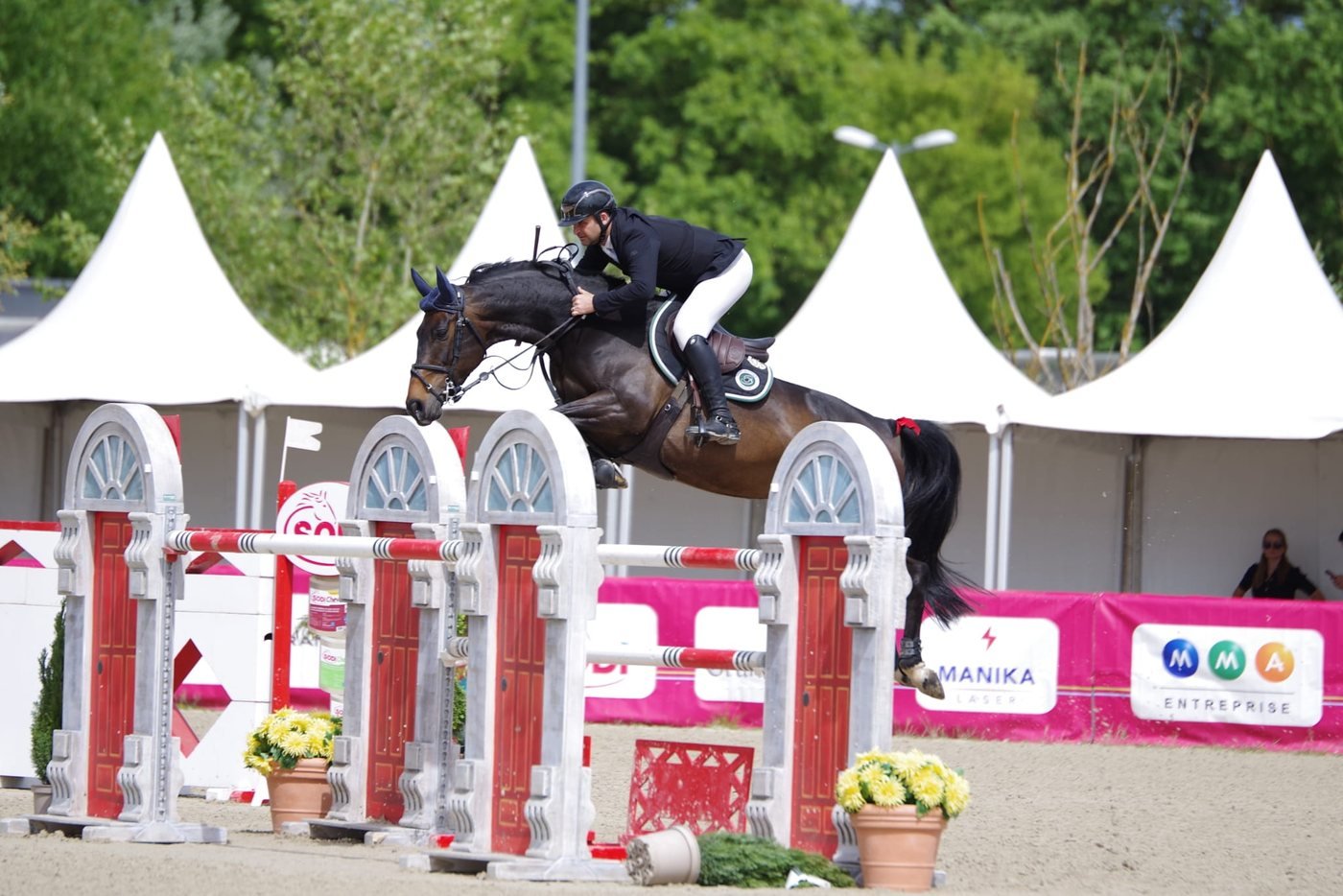 Cavalier en plein saut sur un cheval bai, devant les tentes d'un concours